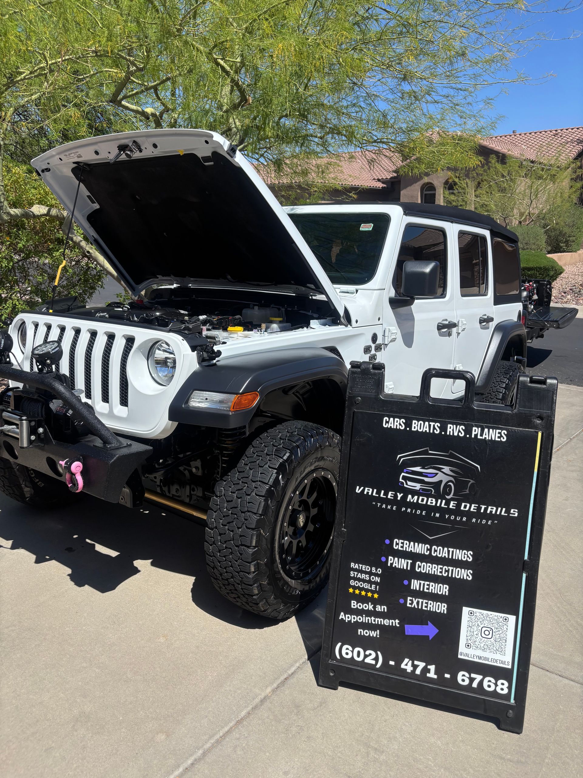 White Jeep Wrangler with hood open beside a black promotional sign on a sunny street