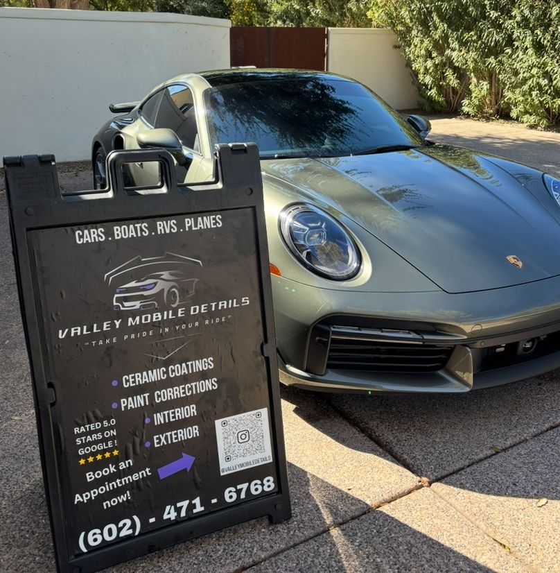 Gray Porsche beside a detailing sign advertising ceramic coating, paint correction, and interior services.