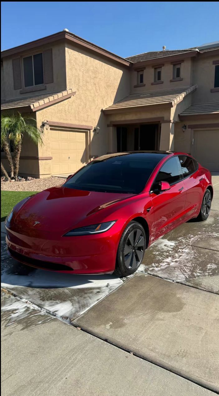 Red Tesla sedan parked in a driveway in front of a tan house on a sunny day