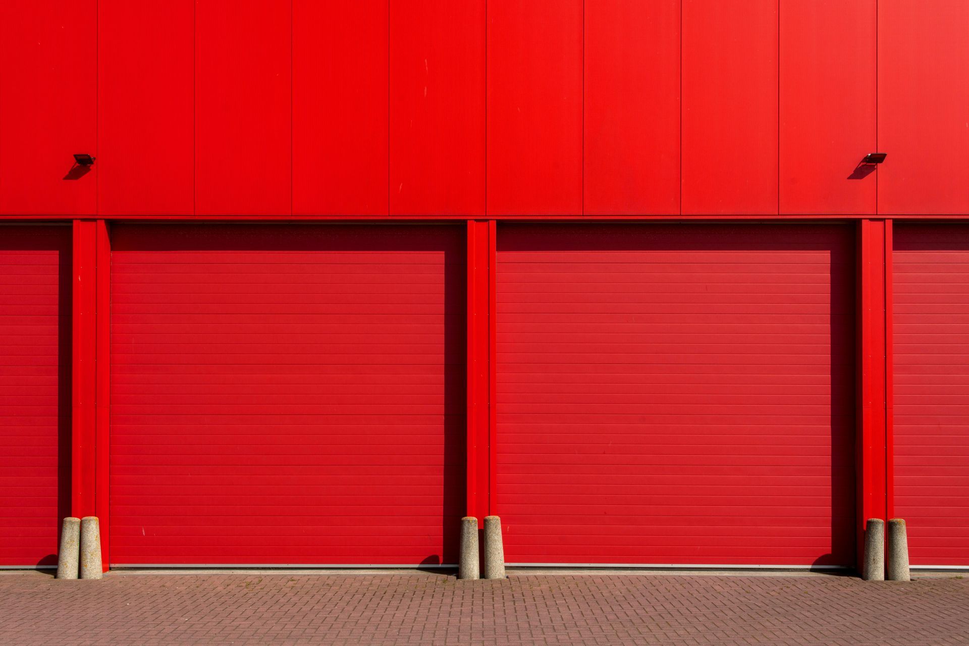 Red industrial building with three closed roller doors and concrete pillars.