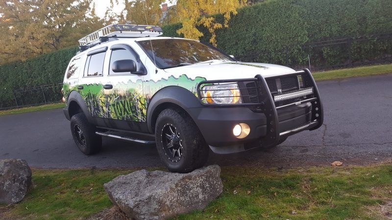 A white SUV with a green forest-themed wrap parked on top of a large rock, equipped with a brush guard and roof rack.