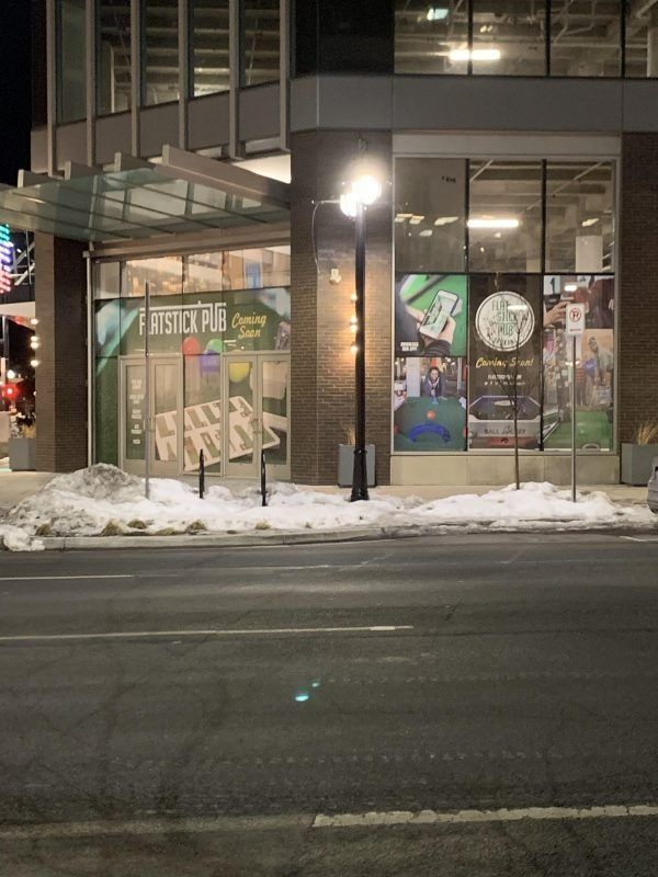A modern building facade at night featuring a Flintlock Pub sign in the window, with piles of snow on the ground.