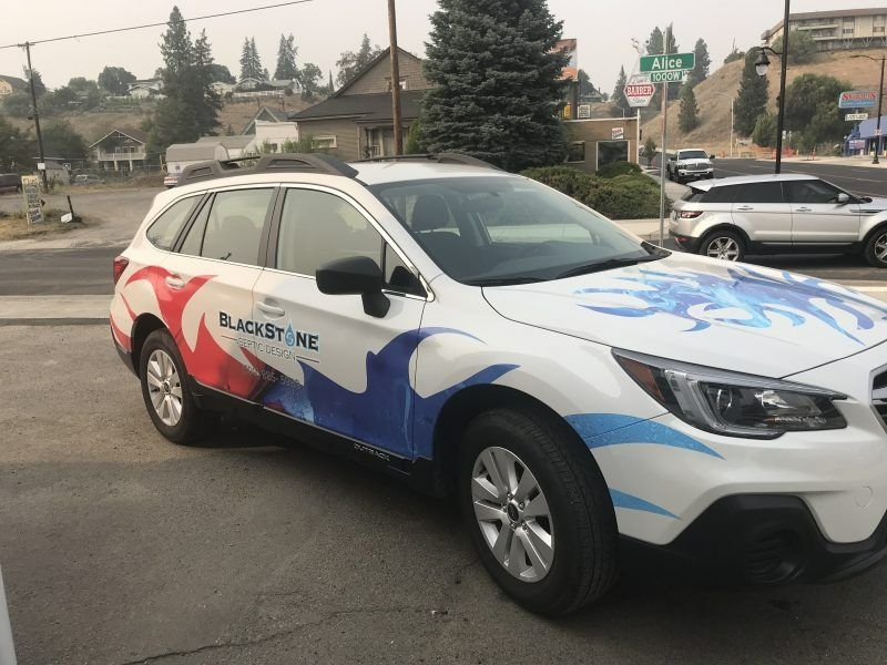 A white Subaru Outback parked in a lot, featuring red and blue swirling vinyl graphics and the Blackstone logo.