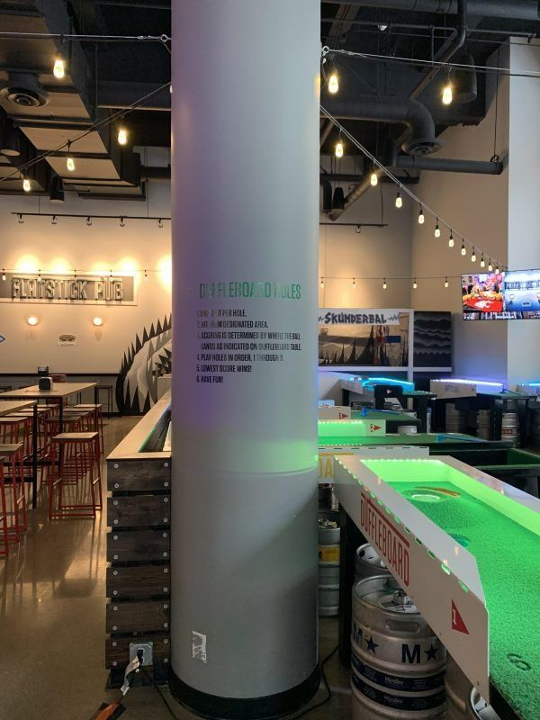 A lounge with string lights features a large pillar with a list of rules and shuffleboard tables with illuminated tracks.