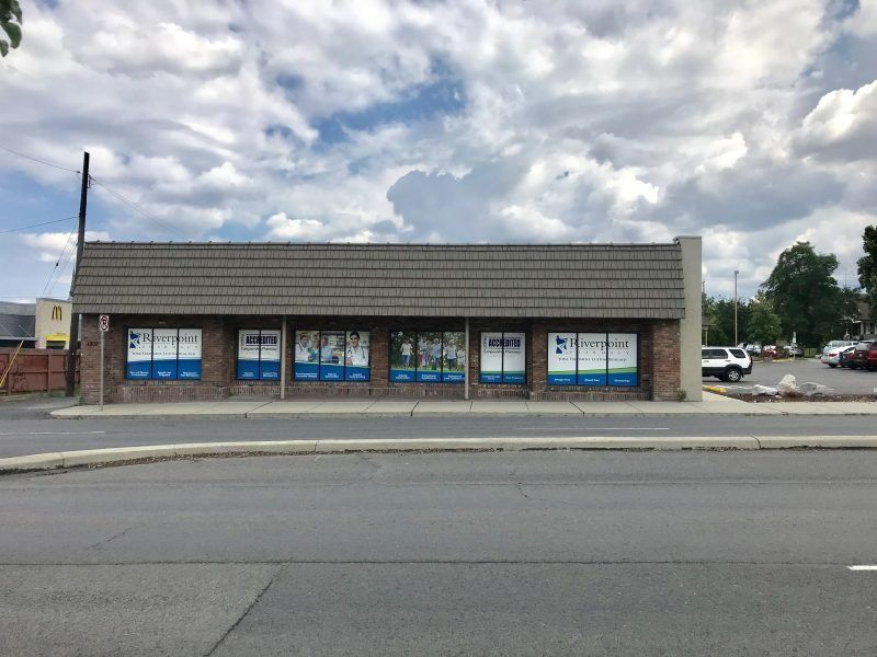 A single-story brown brick commercial building with large glass windows under a dark shingled, sloped roof.