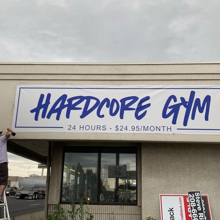 A worker on a ladder installs a banner reading "HARDCORE GYM - 24 HOURS - $24.95/MONTH" on the exterior of a building.