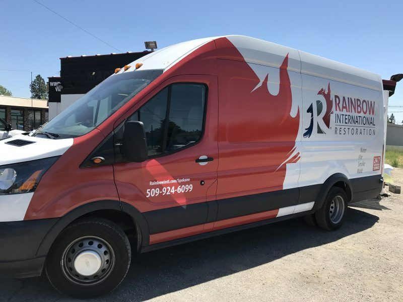 A white and red Rainbow International Restoration van parked on a gravel lot under a clear blue sky.
