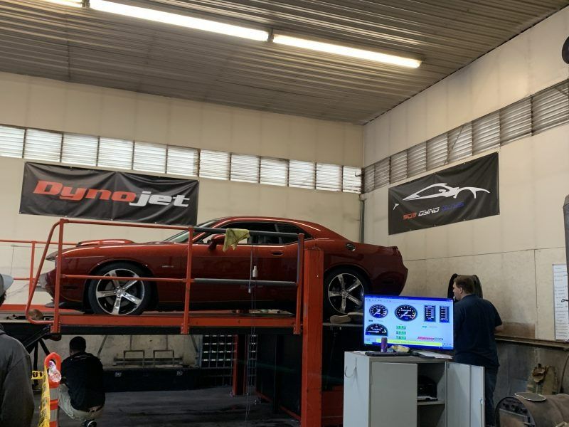 A red muscle car on a dyno lift in an automotive workshop, with two mechanics observing the monitoring screen.