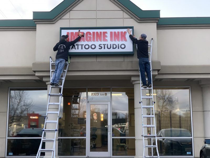 Two people on ladders install a sign for Imagine Ink Tattoo Studio above a storefront entrance.