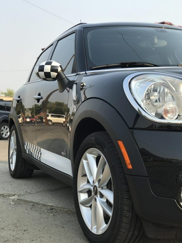A black Mini Cooper with checkered side-view mirrors and white racing stripes parked on an outdoor lot.