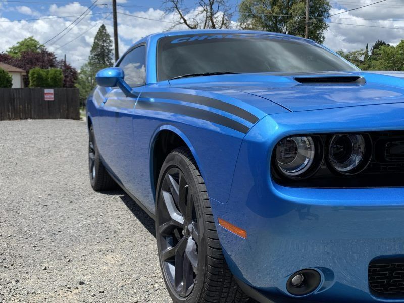 A bright blue Dodge Challenger with black hood stripes, parked on a gravel lot under a cloudy blue sky.