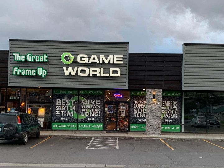 Storefront of "Game World" with a "The Great Frame Up" sign to the left, featuring dark siding and a parking lot in front.