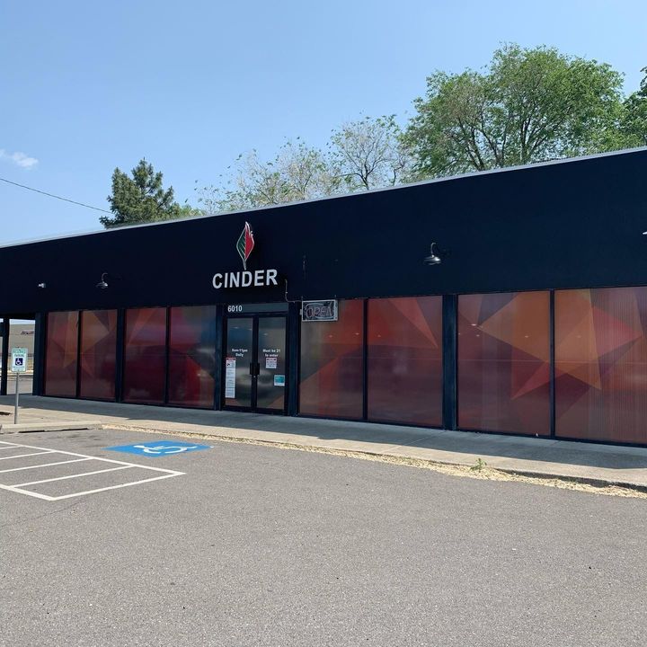 Cinder cannabis storefront with a black facade and orange geometric window graphics, under a bright blue sky.