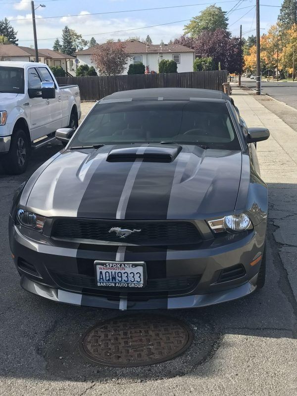 A grey Ford Mustang convertible with black racing stripes parked on a residential street next to a white pickup truck.