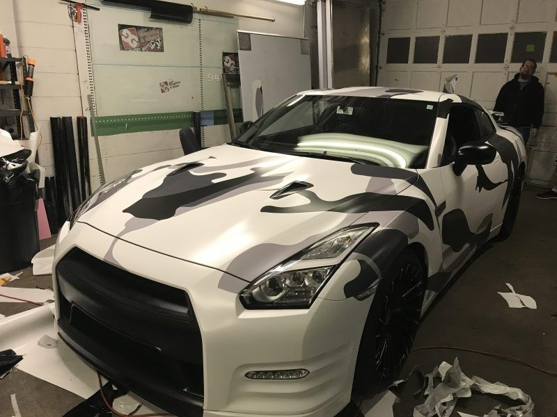 A white Nissan GT-R with a black and gray camouflage vinyl wrap being installed inside a workshop.