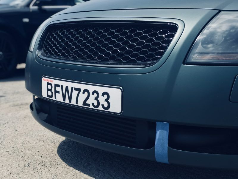 Close-up of a matte dark-green Audi car front featuring a honeycomb grille, license plate BFW7233, and a blue tow strap.