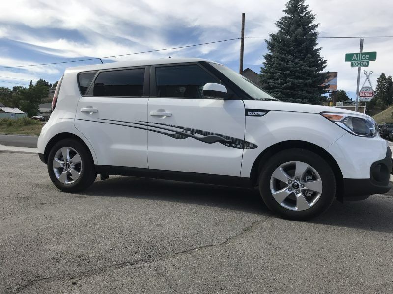 A white Kia Soul parked on pavement with a decorative black vinyl decal along the side, set near a street sign.