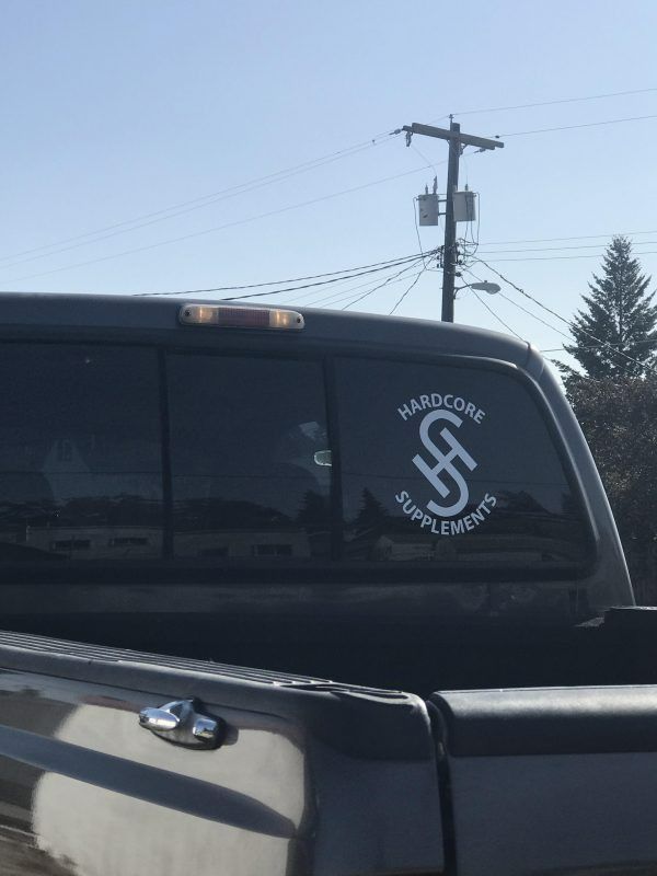 A white decal for Hardcore Supplements on the rear window of a dark pickup truck, parked under a utility pole.