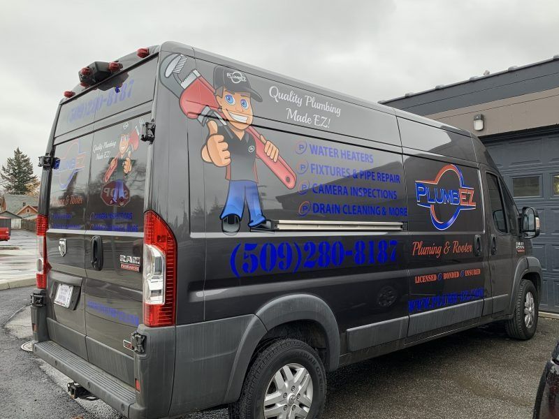 A dark gray plumbing van with a mascot holding a wrench, service list, and phone number printed on the side.