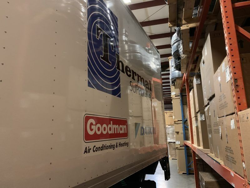 A white truck with Goodman and Daikin logos parked in a warehouse next to industrial shelving filled with boxes.