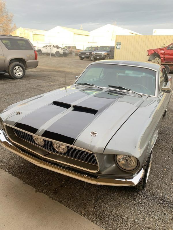 A silver classic Ford Mustang with black racing stripes and hood scoops parked in an outdoor lot.
