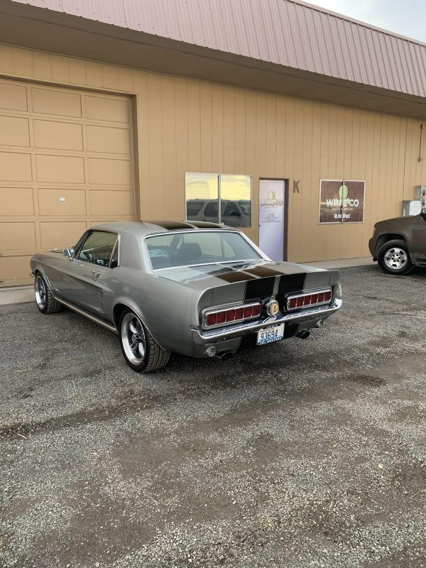 A silver classic Ford Mustang with black racing stripes parked on a gravel lot in front of a tan industrial building.