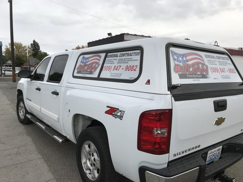A white Chevrolet Silverado pickup truck with a camper shell parked on a street, featuring business branding on the glass.