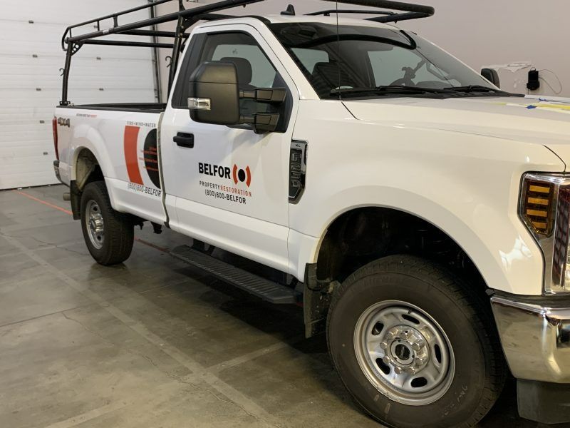 A white Belfor service truck with a utility rack parked inside a warehouse.