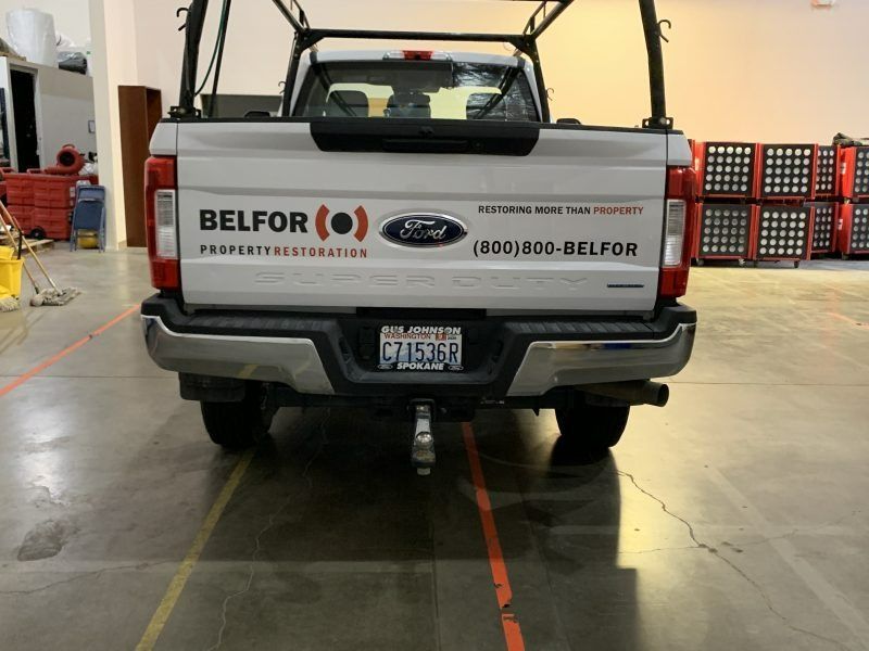 A white Belfor Property Restoration Ford pickup truck parked inside a warehouse with stacked equipment in the background.
