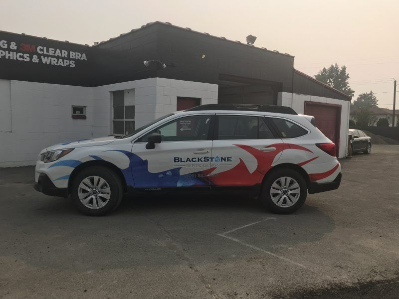 A white Subaru SUV with red and blue graphic decals parked in front of a Blackstone business building.