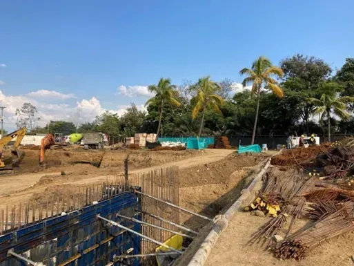 Obra en construcción con cimientos de barras de refuerzo, zanja excavada y palmeras bajo un cielo azul.