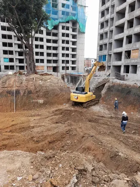Obra en construcción con una excavadora limpiando una fosa de tierra junto a edificios de gran altura sin terminar.