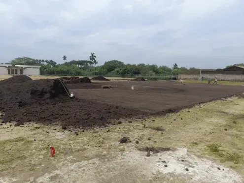 Un campo de tierra con montones de tierra bajo un cielo nublado, bordeado por escasa hierba y edificios a lo lejos.