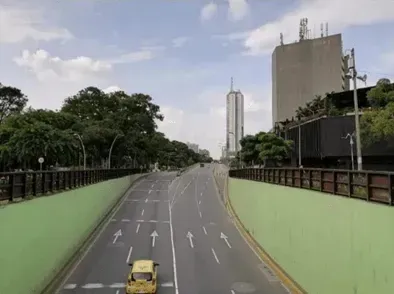 Carretera vacía de varios carriles con muros de contención verdes, árboles y edificios bajo un cielo nublado.