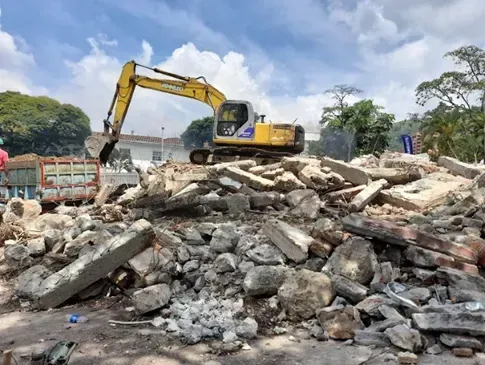Una excavadora retira una gran pila de escombros y hormigón roto en una obra de demolición bajo un cielo nublado.