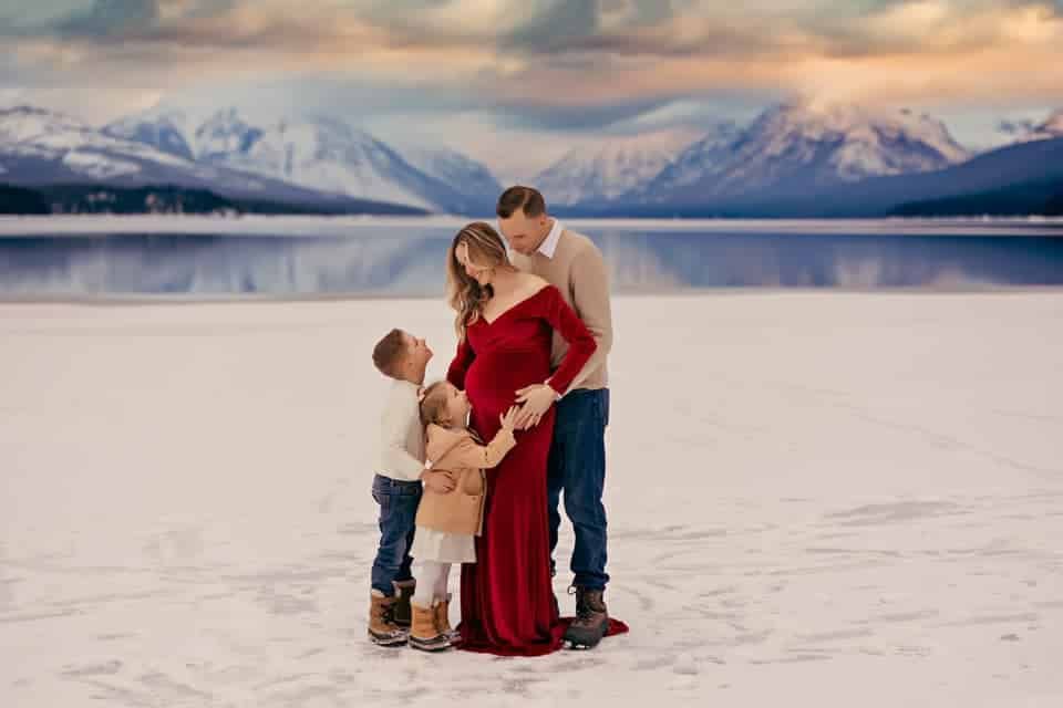 A pregnant woman in a red dress is standing in the snow with her husband and two children.