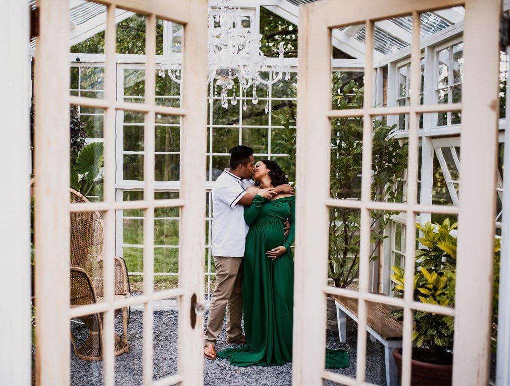 A pregnant woman in a green dress is kissing a man in a greenhouse.