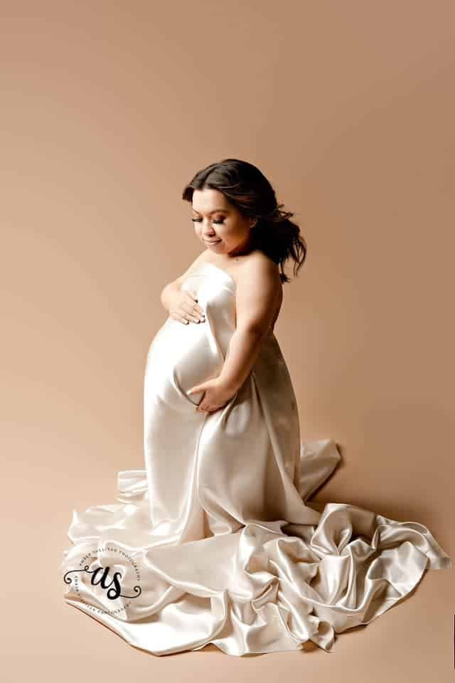 A pregnant woman in a white dress is kneeling down and holding her belly.