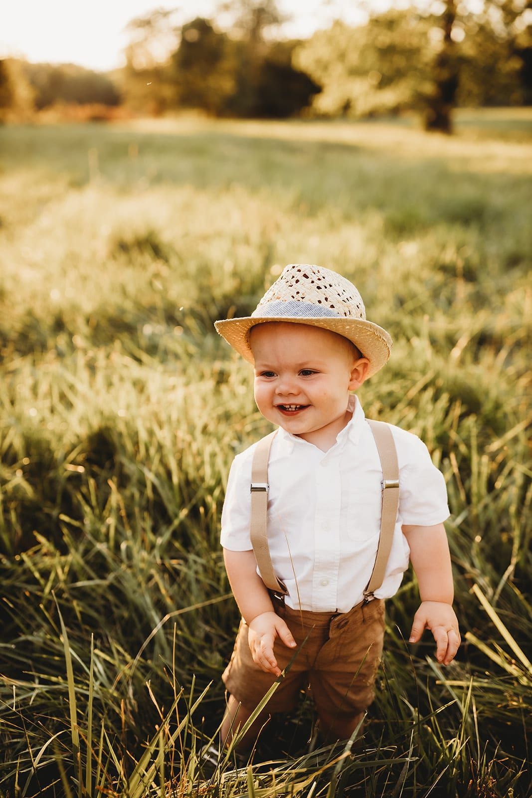 A baby boy wearing a straw hat and suspenders is standing in a field.