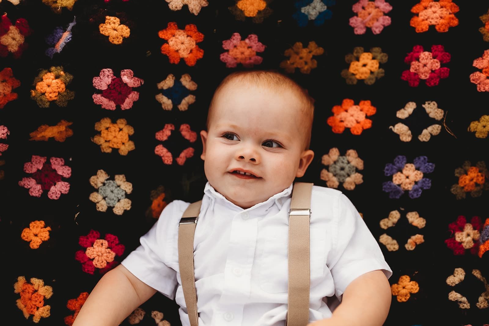 A baby wearing suspenders and a white shirt is sitting in front of a blanket with flowers on it.
