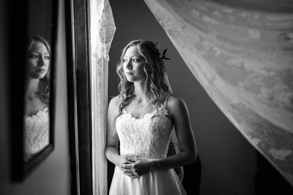 A black and white photo of a bride in a wedding dress looking out of a window.