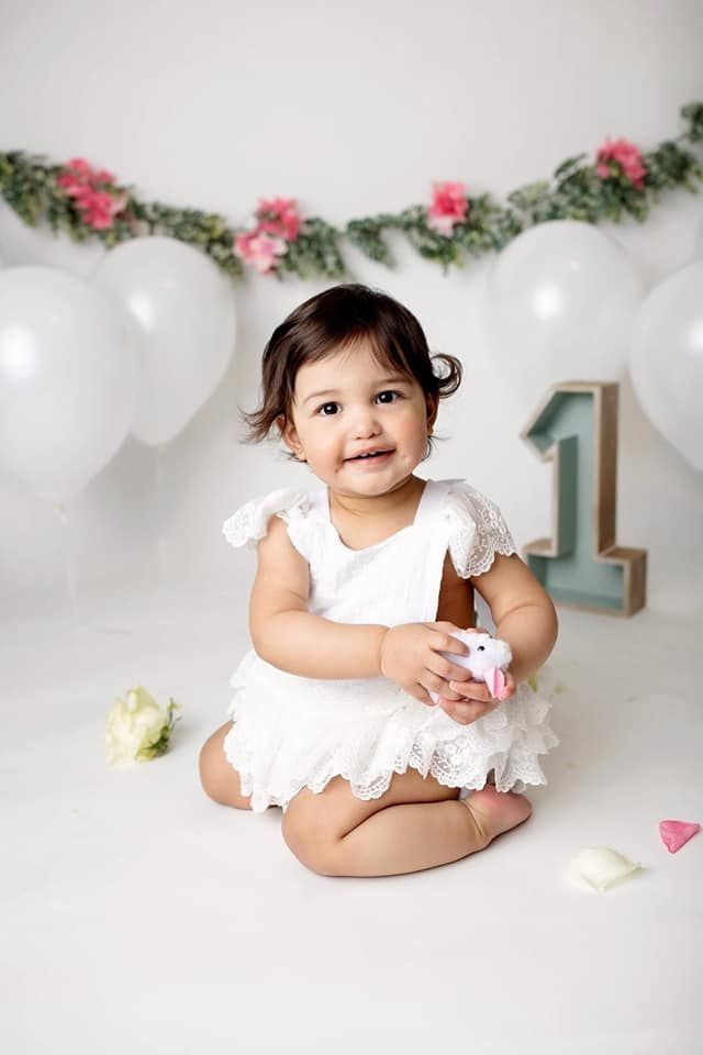 A baby girl in a white dress is sitting on the floor holding a cupcake.