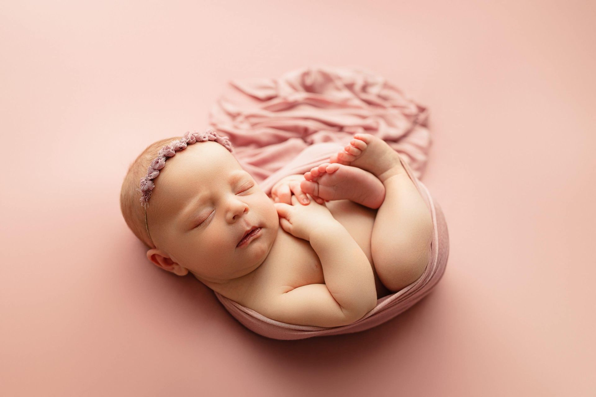 A newborn baby is sleeping in a pink bowl on a pink background.