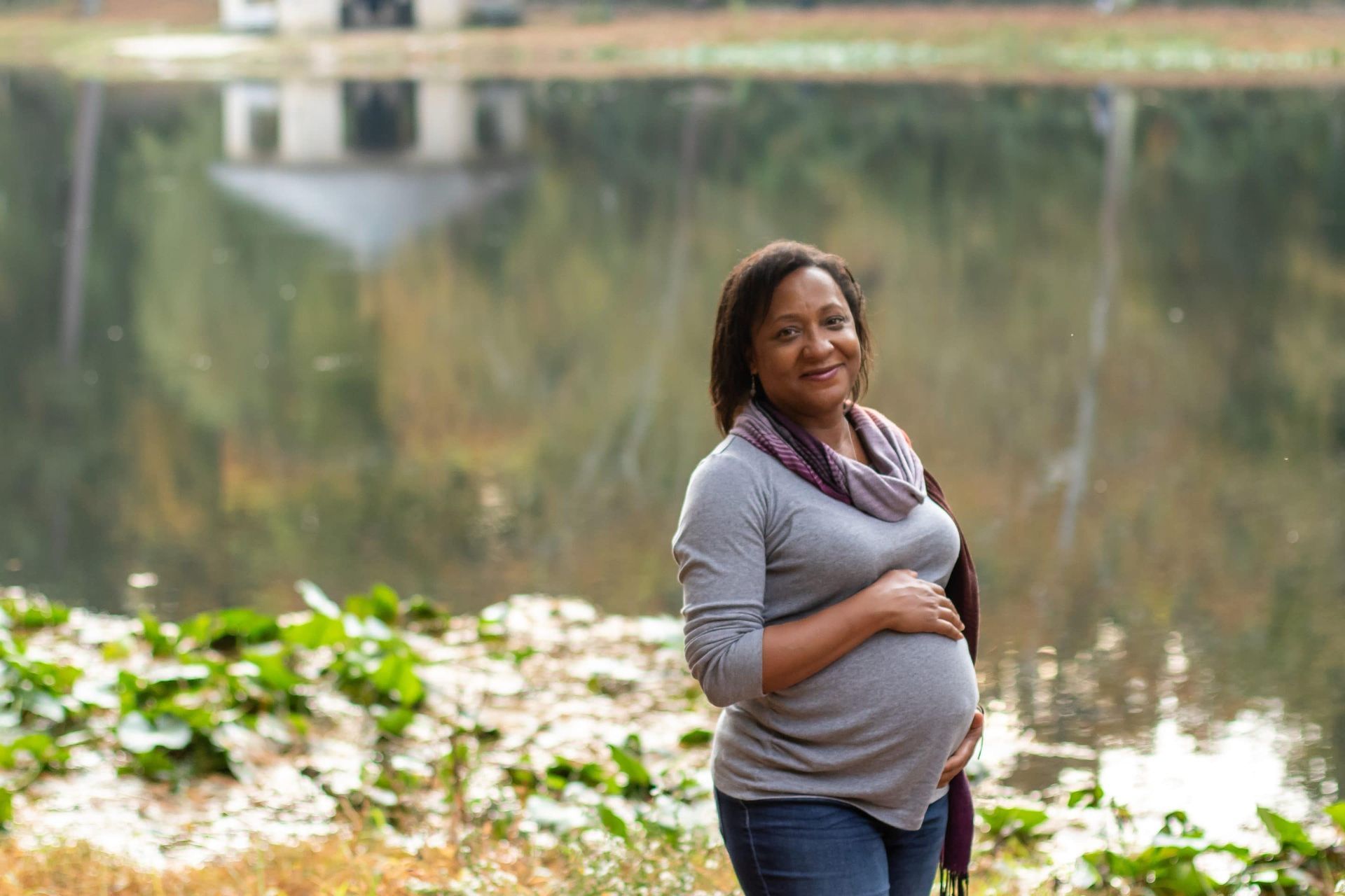 A pregnant woman is standing in front of a lake holding her belly.