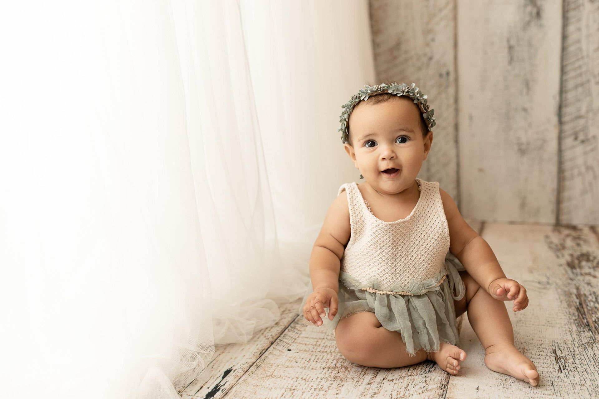 A baby girl is sitting on a wooden floor wearing a flower crown.