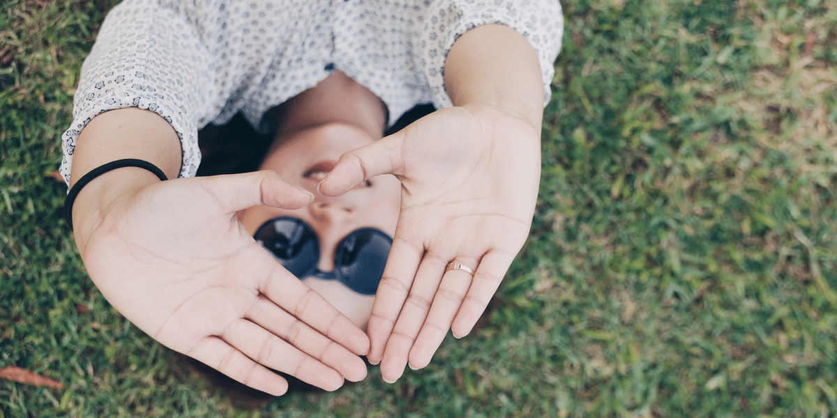 A woman is laying on the grass making a heart shape with her hands.