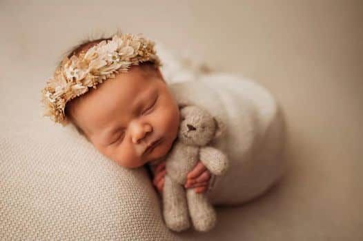 The baby is wearing a flower crown and holding a teddy bear.