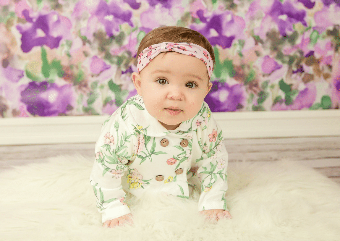 A baby girl is crawling on the floor in front of a purple floral background.