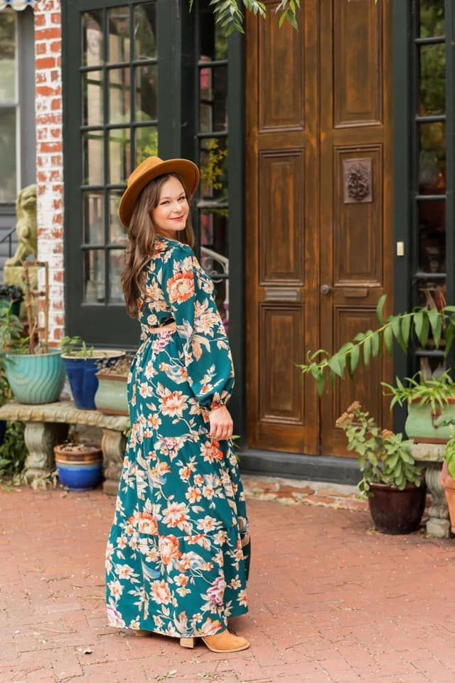 A woman in a floral dress and hat is standing in front of a door.