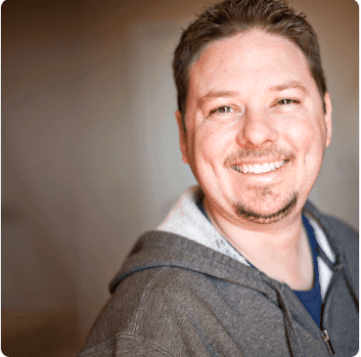 A man with a beard wearing a grey hoodie smiles for the camera